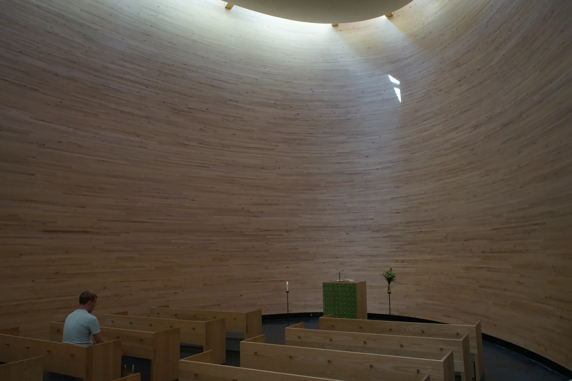 A lone figure sits in a curved wooden chapel, light falling from an opening above.